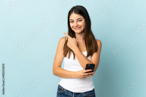 Young caucasian woman using mobile phone isolated on blue background pointing to the side to present a product