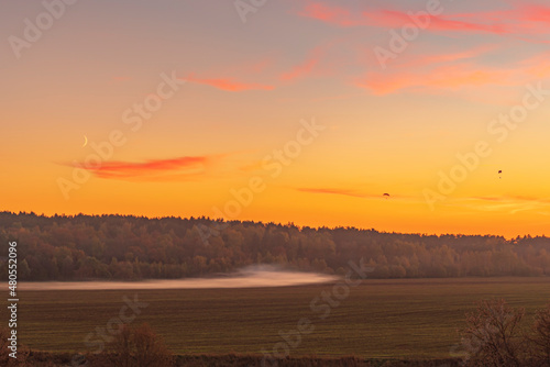 Sunset in the mist field with silhouette of paragliders flying over the orange sky