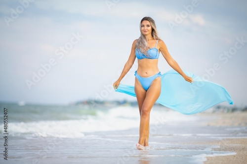 A girl with blond hair in a bluish swimsuit and a bright shawl walks along the beach