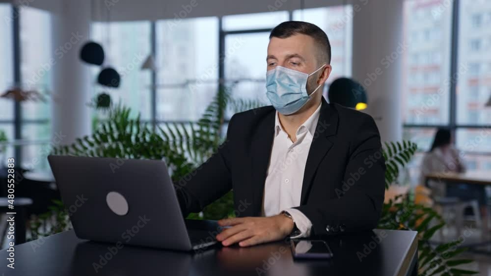Male employee wearing suit and mask sitting in front of laptop. Man following pandemic restrictions during work in the office. Blurred background. Male employee wearing suit and mask sitting in front of laptop. Man following pandemic restrictions during work in the office. Blurred background.