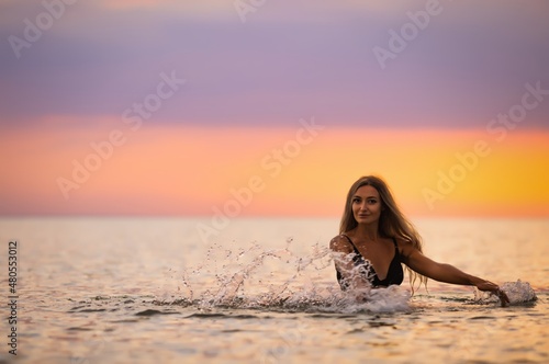A girl with blond hair in a black swimsuit splashes to the sides while sitting in an estuary on a sunset background © YouraPechkin