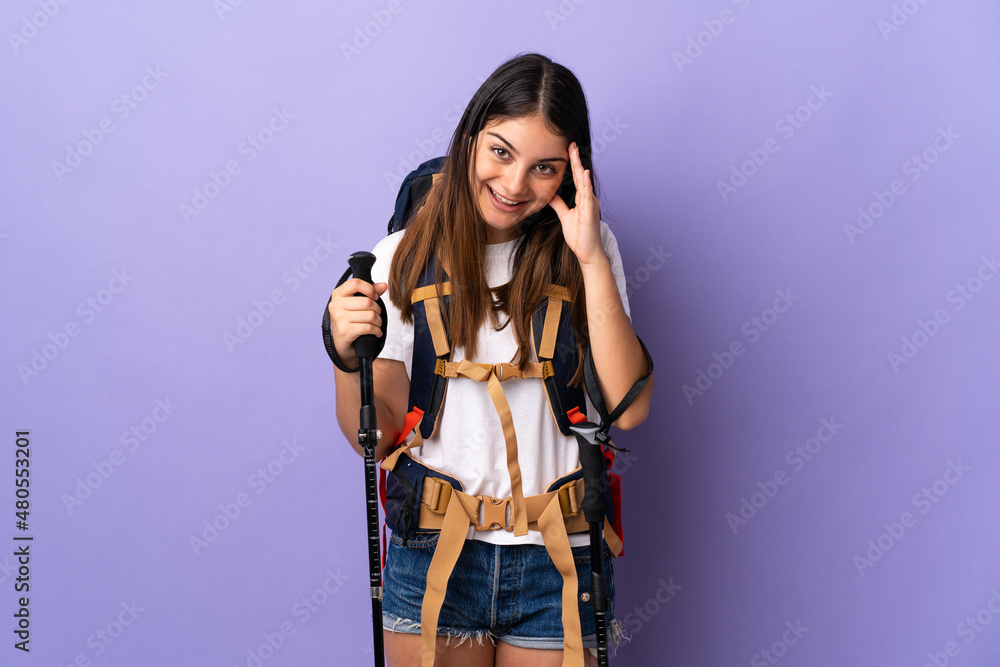 Young woman with backpack and trekking poles isolated on purple background with surprise expression Young woman with backpack and trekking poles isolated on purple background with surprise expression