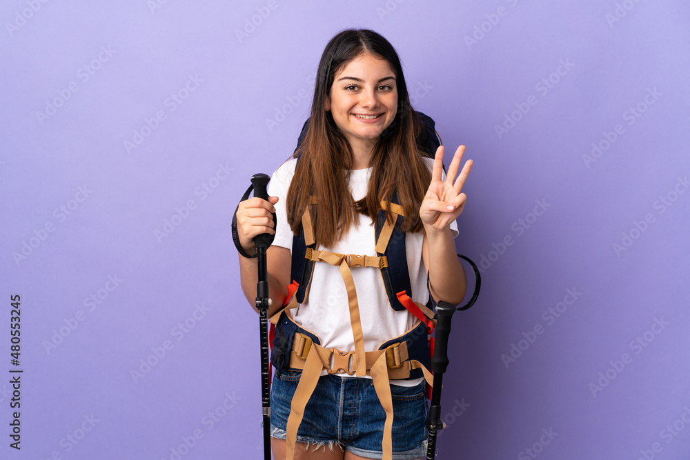 Young woman with backpack and trekking poles isolated on purple background happy and counting three with fingers Young woman with backpack and trekking poles isolated on purple background happy and counting three with fingers