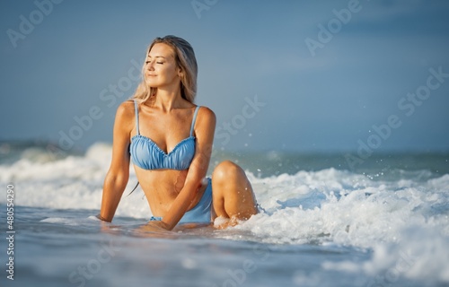 A girl with blond hair in a delicate swimsuit sits by the sea  enjoying the splash of waves