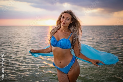 A girl in a blue swimsuit and a bright pareo posing against the background of a sunset in estuary with transparent water