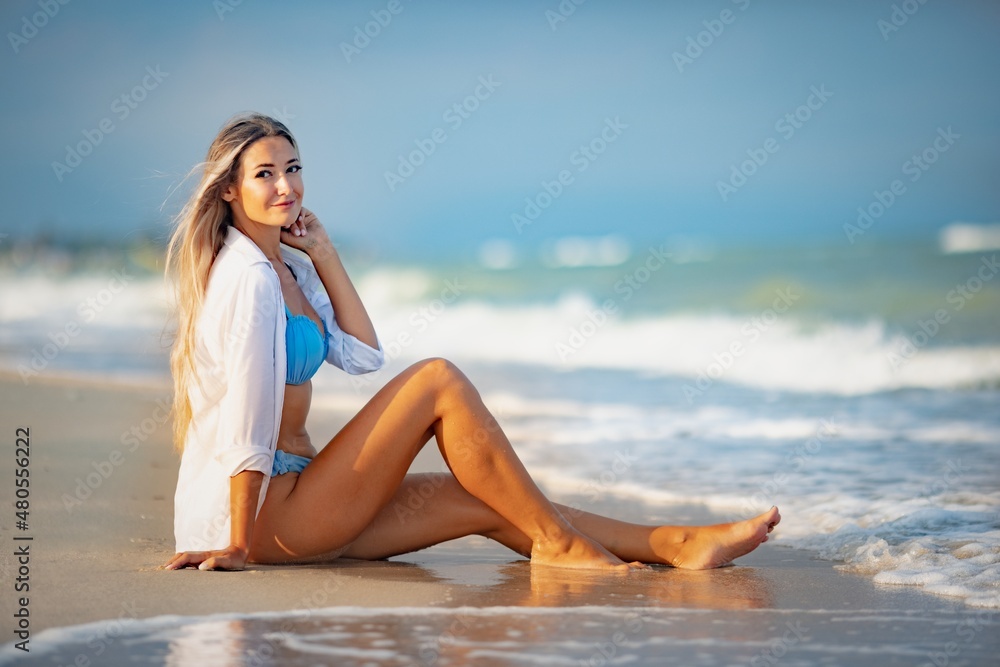 A girl in a bluish swimsuit and shirt sits near the water edge and peers into the horizon