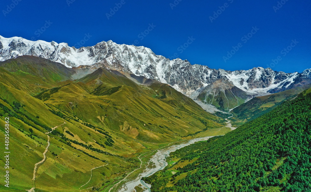 Aerial view of a snow-capped mountain ridge on a summer sunny day against a clear blue sky background. Shkhara Glacier in the central part of the Greater Caucasus Range in Georgia.