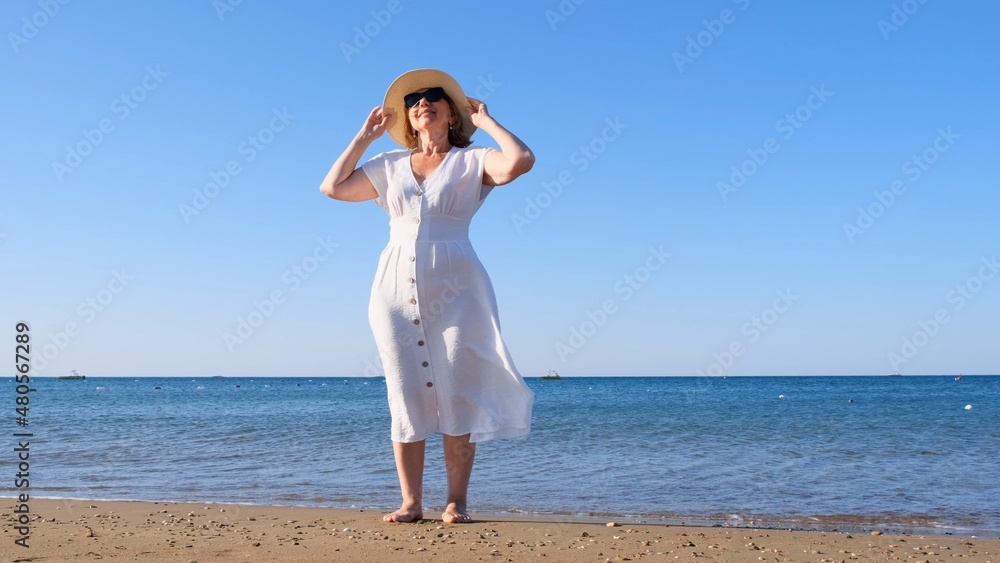 mature woman in sunglasses in a straw hat and white dress walks along the blue sea coast on a sunny summer day, enjoying freedom and relaxation. The concept of a senior citizen everyday life mature woman in sunglasses in a straw hat and white dress walks along the blue sea coast on a sunny summer day, enjoying freedom and relaxation. The concept of a senior citizen everyday life