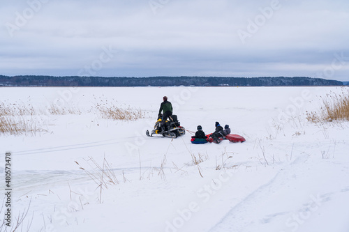 Winter landscape of a large lake covered with ice and snow. You can see the figure of a man on a snowmobile, which rolls several children on tubing.