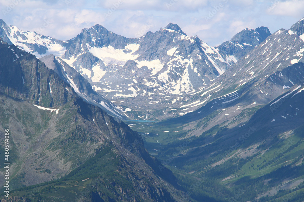 Rocky peaks of a high-mountain ridge on the Kara-Turek pass in Altai with snow, a glacier and a small lake in summer, rocky mountain slopes on the right and left, a sky with clouds, sunny