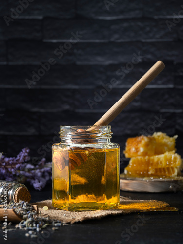 A glass jar with transparent golden honey on a dark table. Honeycombs on a wooden board and lavender twigs in the background. Dark brick background