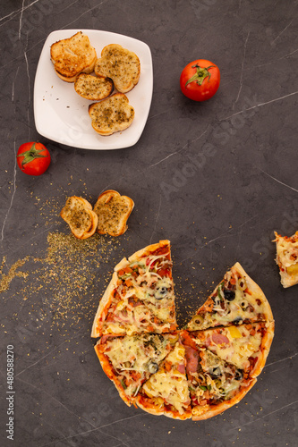 elevated perspective of a dark table with a plate of garlic bread with oregano, sliced pizza with cheese, pineapple and ham, fast food, traditional and handmade in studio