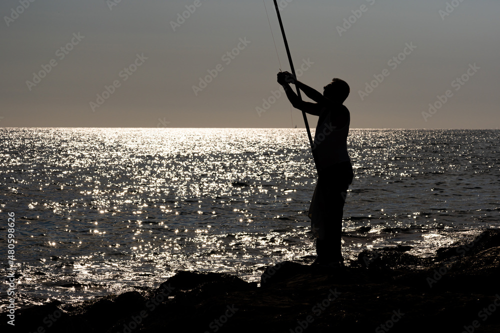 Backlit fisherman on a beach breakwater.