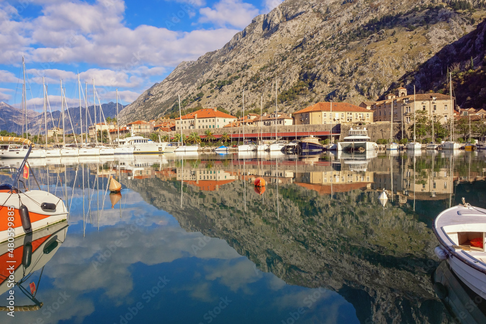 View of marina near Old Town of Kotor on sunny winter day.  Mountains, boats  and walls of Kotor Old Town are reflected in water. Montenegro. Beautiful Mediterranean landscape
