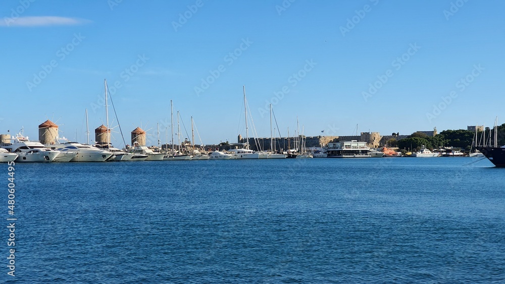 In sunny weather, yachts are moored in a marina with windmills on the island of Rhodes. Part of the fortress wall is visible, view from the sea. The marina is calm. The best marinas in Rhodes. No wind