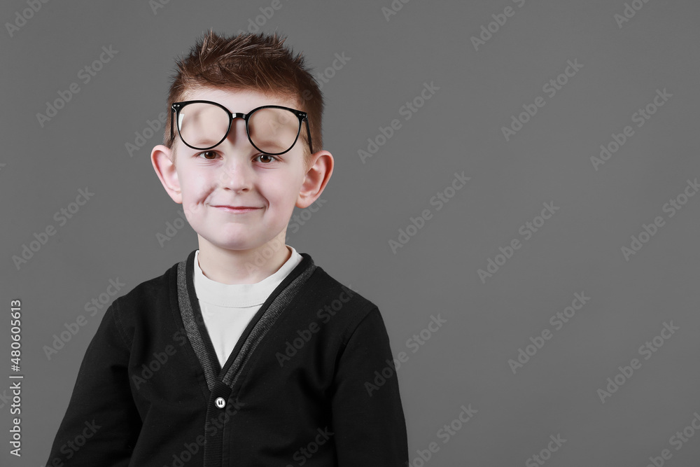 Smart child boy adjusts his glasses and having fun on the grey background, close-up. Studio juvenile portrait in casual clothes. copy space
