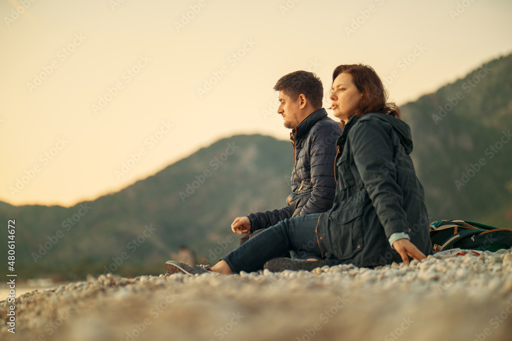 A man and a woman sit on a stone beach and enjoy the winter sea and a beautiful sunset. Date. Romance. Husband and wife. Tourism. Travelers. Vacation. A day off in nature.