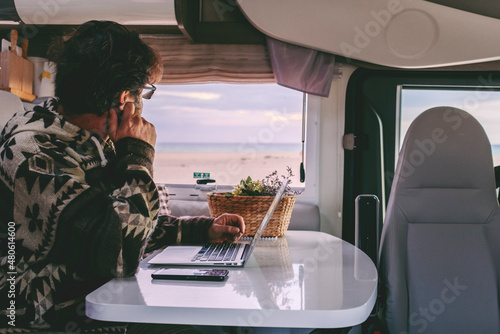 Man working inside camper van sitting at the table and looking outside the window the beautiful beach and ocean in background. Concept of freedom lifestyle and remote online worker people