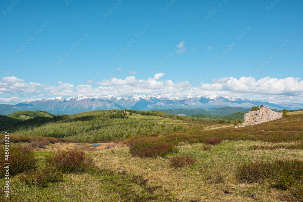 Fototapeta premium Atmospheric landscape with big rock on hill with view to sunlit mountain vastness and high snowy mountain range under low clouds line on horizon. Impressive scenery with vast mountains in sunlight.