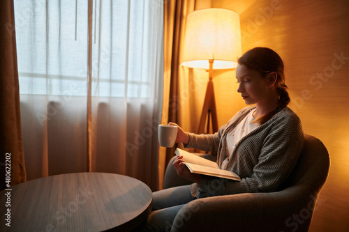 A young blonde woman sits in an armchair with a cup of tea, coffee and reads a book. Near the window, a floor lamp illuminates the room with orange light. Rest, home comfort.