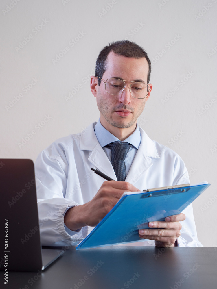Young doctor man with eyeglasses writes on clipboard for his work, doctor sitting in front of his desk with laptop Young doctor man with eyeglasses writes on clipboard for his work, doctor sitting in front of his desk with laptop