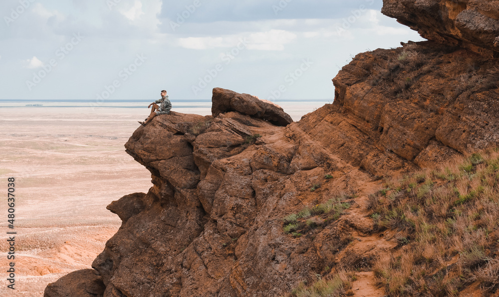 Man from the side, sitting on top of a rock, looking to distance beyond the horizon.Red mountain Big Bogdo near Lake Baskunchak. Astrakhan region of Russia. Martian landscape