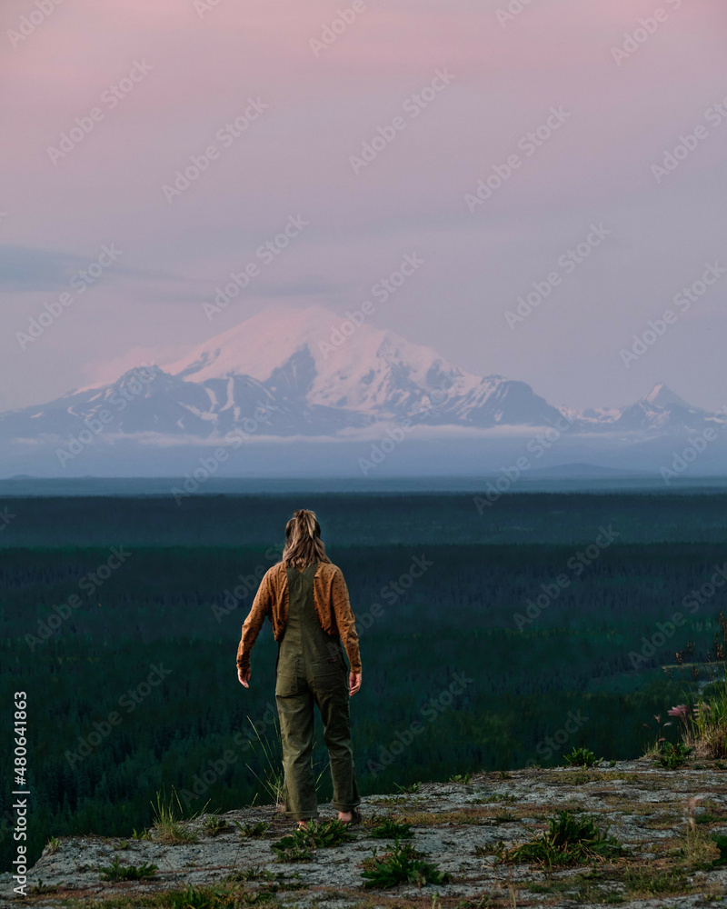 Adventure girl hiking in Alaska with Volcano Mountain in background during purple red and orange sunset with pink sky and alpenglow on peak