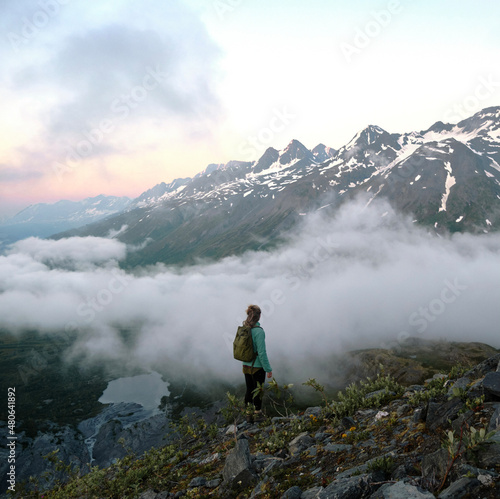  Adventure girl with backpack hiking on mountains during sunset above clouds in Alaska with colorful sky and lake in valley below