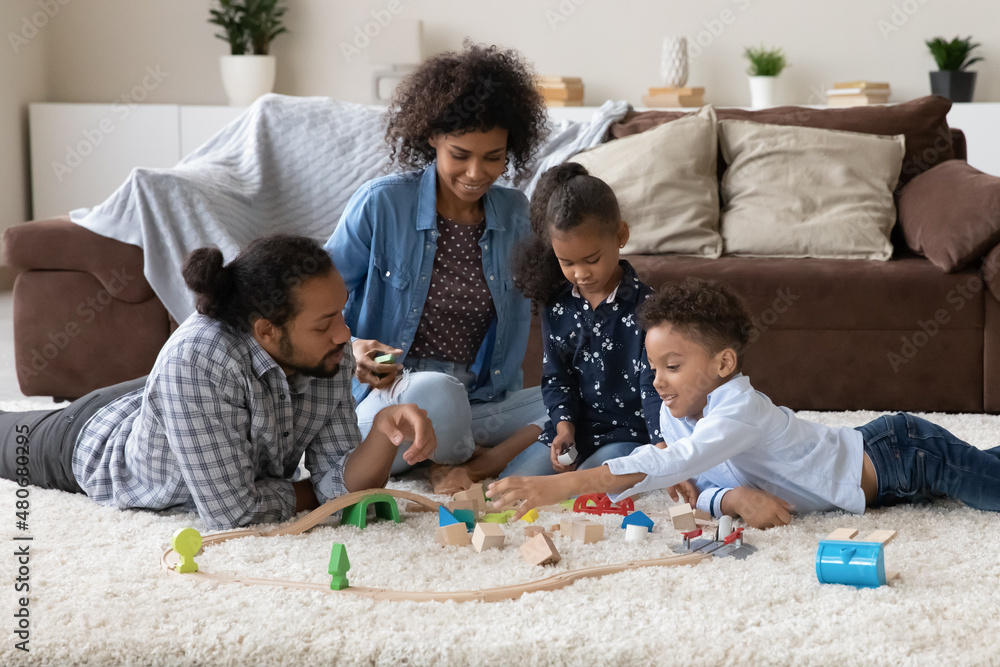 Happy Black couple of parents helping two kids to build toy road model, playing with wooden construction blocks on warm soft carpeted floor, enjoying creative game, hobby, family leisure time Happy Black couple of parents helping two kids to build toy road model, playing with wooden construction blocks on warm soft carpeted floor, enjoying creative game, hobby, family leisure time