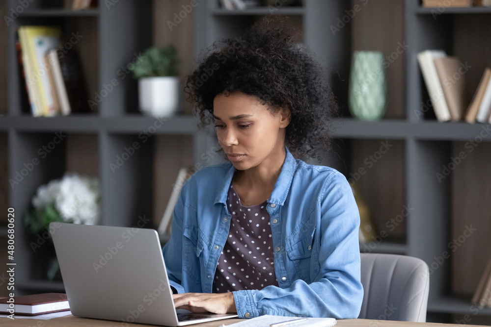 Serious young African business woman in casual using laptop, working at home office workplace. Student girl, remote employee chatting online on computer, using internet app, watching webinar Serious young African business woman in casual using laptop, working at home office workplace. Student girl, remote employee chatting online on computer, using internet app, watching webinar