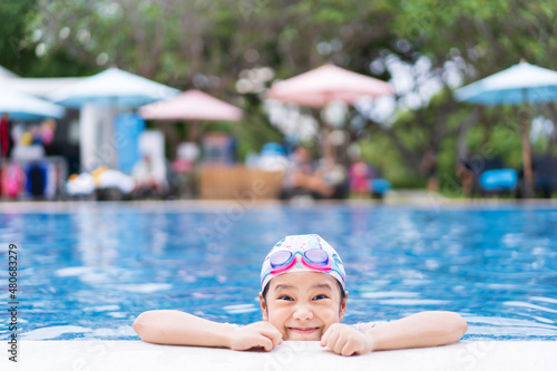 Asian child or kid girl wearing swimsuit and cap with goggles on edge swimming pool and smile with happy fun in waterpark for learning swim to exercise on summer school or vacation travel at hotel © kornnphoto