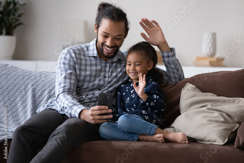 Happy Afro American millennial dad and cheerful cute kid talking on video call together, waving hand hello at smartphone screen, using telephone for online conference. Family communication concept © fizkes