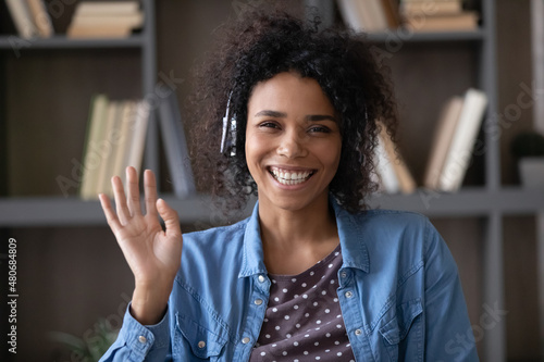 Happy excited African student, employee in wireless headphones waving hand hello with toothy smile, laughing at camera, making video call at home. Black woman talking online head shot portrait screen