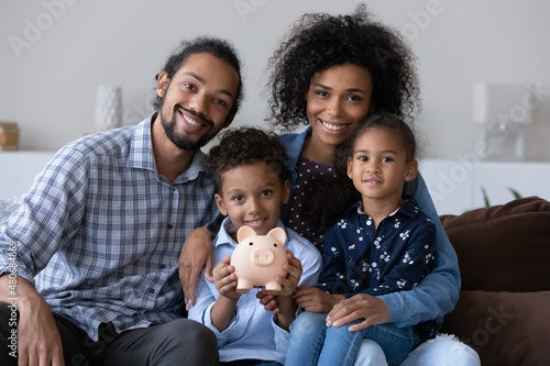 Fototapeta Happy Black family couple and little kids making financial reserve, donation funk, keeping money in piggybank, showing moneybox, looking at camera, smiling