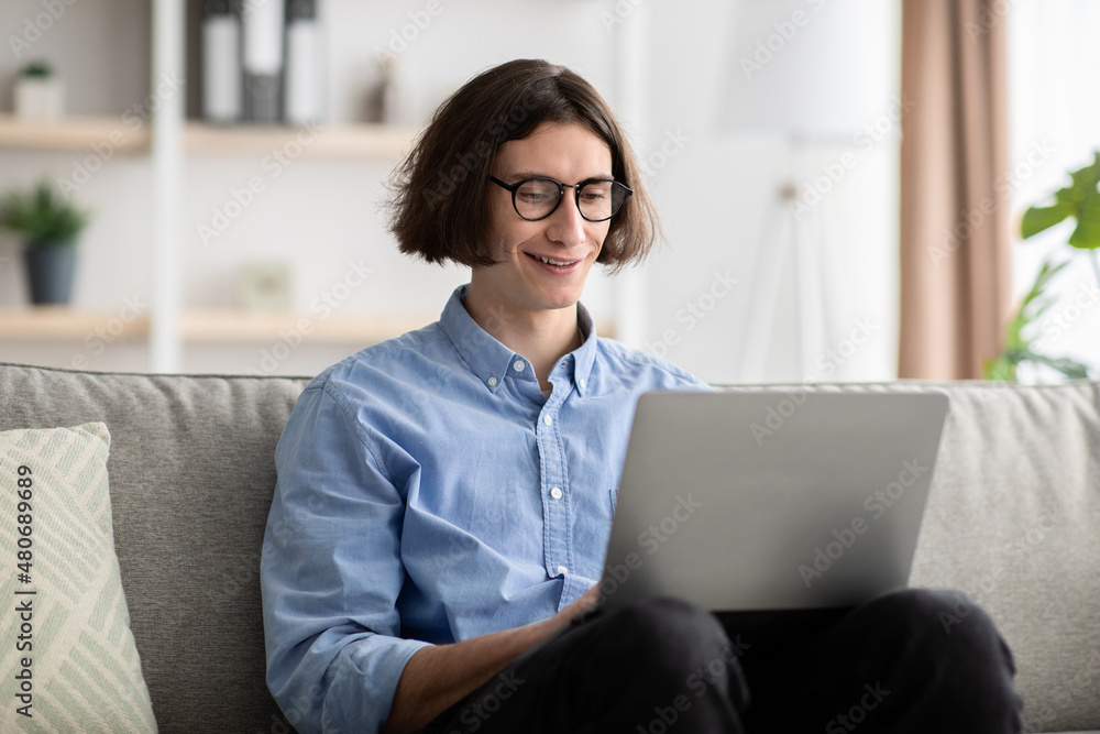Distant work. Happy young freelancer man using laptop computer, working remotely frm home, sitting on sofa Distant work. Happy young freelancer man using laptop computer, working remotely frm home, sitting on sofa