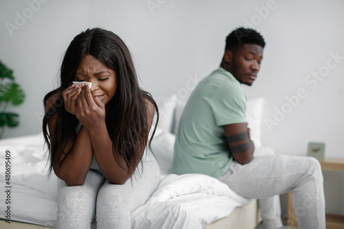 Unhappy sad young african american woman crying, ignoring her husband on bed in bedroom interior