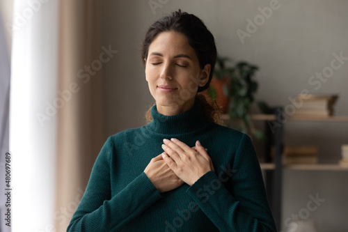 Head shot peaceful young attractive woman holding folded hands on chest, praying waiting for miracle, feeling thankful indoors. Sincere happy female volunteer showing kindness or expressing gratitude. photo
