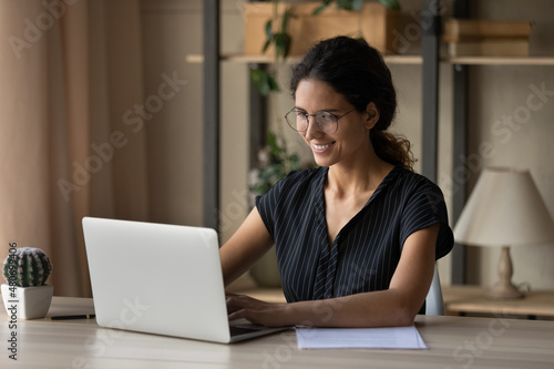 Fotografie Happy attractive millennial hispanic latina businesswoman in eyeglasses working on computer in modern home office, communicating distantly with partner or colleague, consulting client remotely