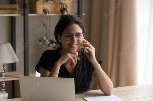 Smiling young pretty hispanic latina business lady in eyeglasses holding distant phone call, working distantly on computer in modern home office. Happy millennial employee discussing project remotely.
