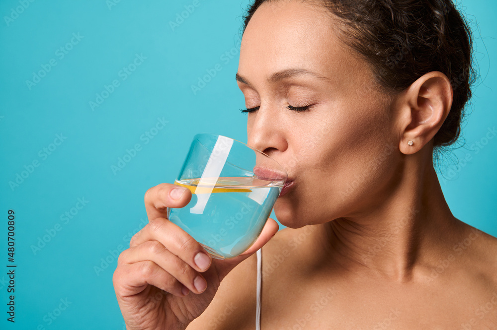 Headshot of a beautiful African woman with fresh healthy skin drinking lemon water from transparent glass, isolated over bright blue background with copy ad space. Healthy eating and lifestyle concept Headshot of a beautiful African woman with fresh healthy skin drinking lemon water from transparent glass, isolated over bright blue background with copy ad space. Healthy eating and lifestyle concept
