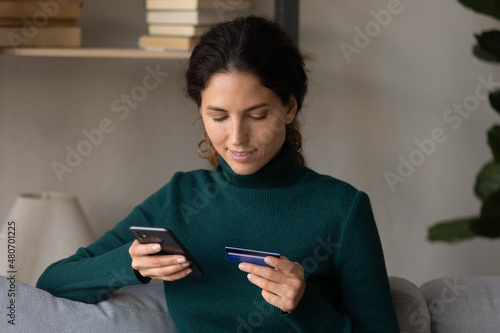 Smiling young hispanic latin caucasian woman entering payment information from bank card in mobile shopping app, transferring money, paying for services online, purchasing goods in internet store.