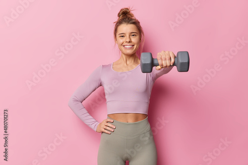 Sporty young woman being in good physical shape keeps hand on waist raises dumbbell has regular fitness training wears sportsclothes poses against pink background. Gym workout and sport concept