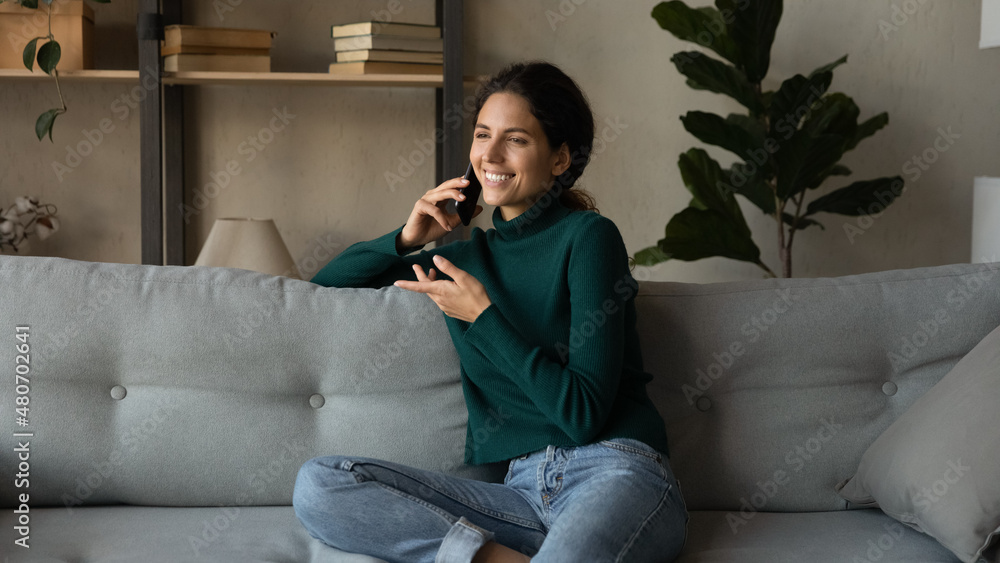 Fototapeta premium Smiling beautiful young woman involved in mobile phone call conversation with friends, talking chatting speaking communicating distantly resting on comfortable sofa alone in modern living room.