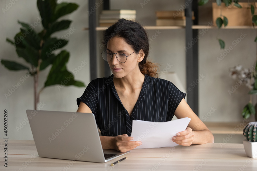 Concentrated smart young hispanic caucasian business lady employee in eyewear analyzing paper report, developing online project growth strategy, review sales data statistics in modern home office. Concentrated smart young hispanic caucasian business lady employee in eyewear analyzing paper report, developing online project growth strategy, review sales data statistics in modern home office.