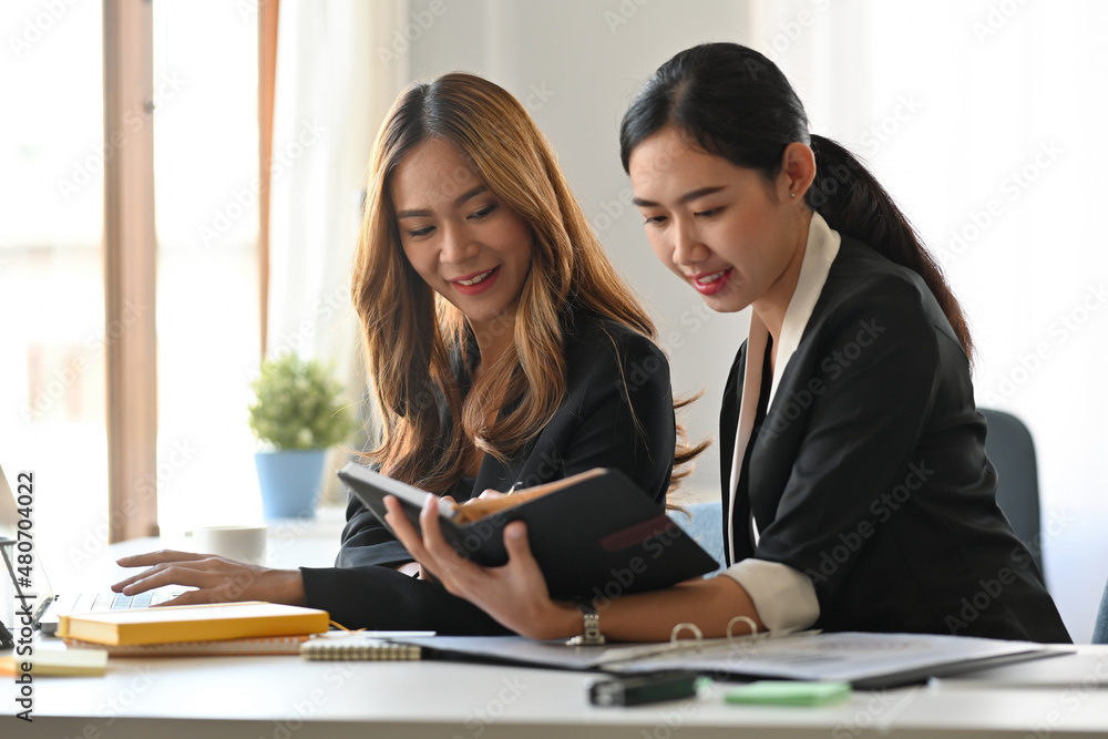 Photo of a young businesswoman and her secretary working together at the wooden working desk surrounded by a computer laptop and business document. Photo of a young businesswoman and her secretary working together at the wooden working desk surrounded by a computer laptop and business document.