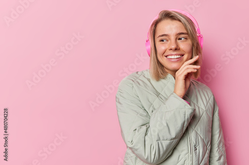 Happy tender young woman with bob hair touches face smiles broady looks away happily listens music via headphones wears jacket isolated over pink background blank copy space for your promotion photo