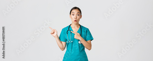 Covid-19, healthcare workers, pandemic concept. Impressed and amazed female asian nurse in scrubs asking question with intrigued expression. Doctor looking thrilled and pointing upper left corner