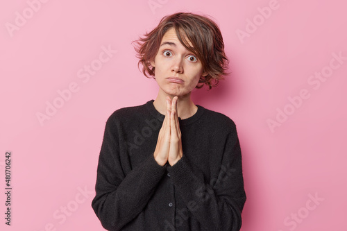 Fotografía Coy beautiful woman with short hair keeps hands in pray beg gesture says please wears casual black jumper isolated over pink background asks for favor needs something