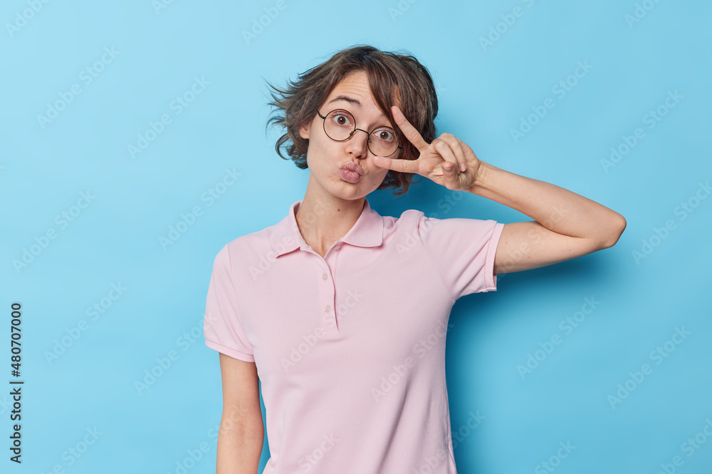 Beautiful young woman with short dark hair keeps lips folded makes peace gesture over eye wears round spectacles for good vision and casual t shirt isolated over blue background shows salute