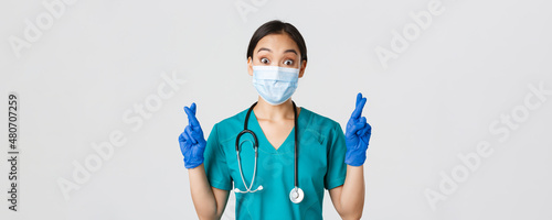 Covid-19, coronavirus disease, healthcare workers concept. Excited and hopeful asian female doctor, nurse in medical mask and rubber gloves cross fingers good luck, makign wish, white background photo